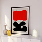The Sundown print framed and propped on a white sideboard in a serene interior, with soft lighting that enhances the striking contrast of the red and black forms.

