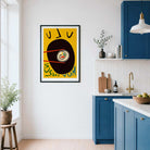 A framed abstract art print featuring a yellow background, a large black circle, and stylized text hangs on a white wall in a modern kitchen with blue cabinets.