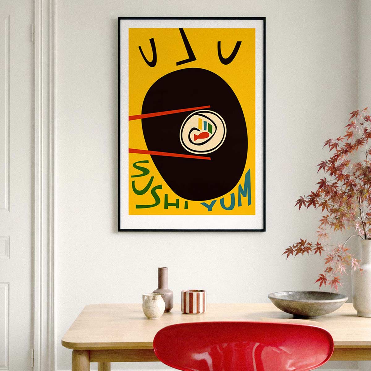A framed abstract sushi-themed art print hangs on a white wall above a wooden table with decorative vases, a bowl, and a red chair. The print features a yellow background with a large black oval, red chopsticks, and stylized sushi and Japanese characters.