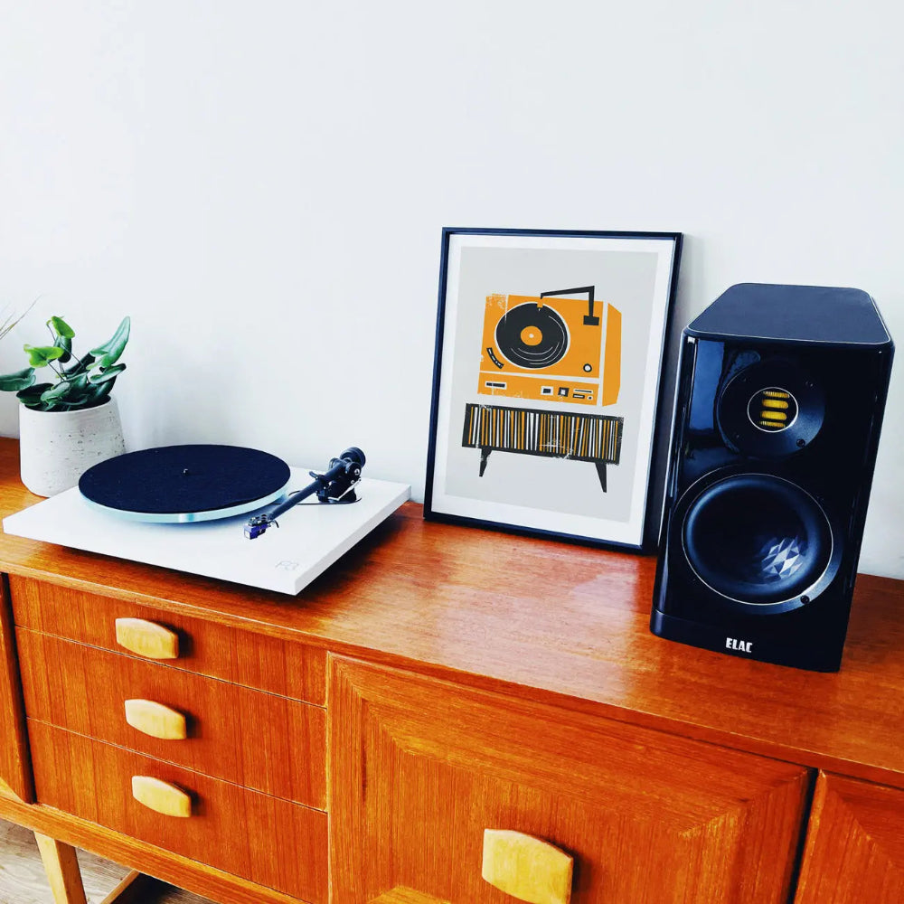 Framed retro record player print displayed on a wooden sideboard beside a modern speaker setup—where music nostalgia meets contemporary living.
