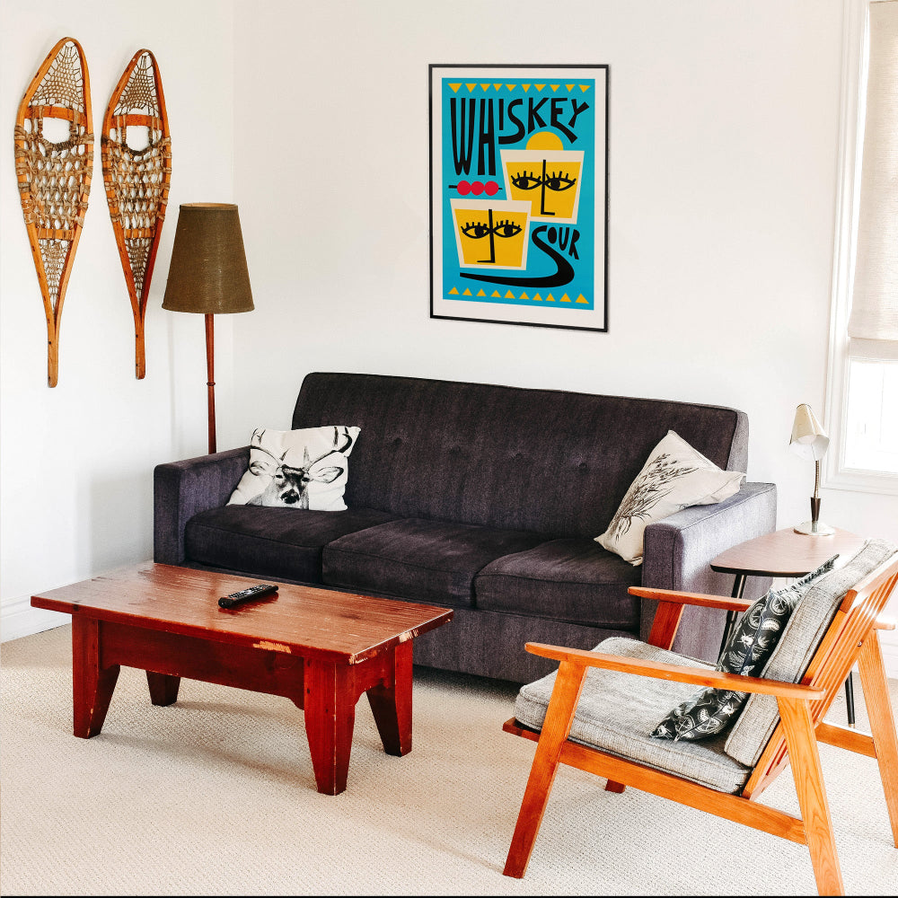 Framed Whiskey Sour cocktail print hanging above dark sofa in a retro-style living room with warm wood furniture.