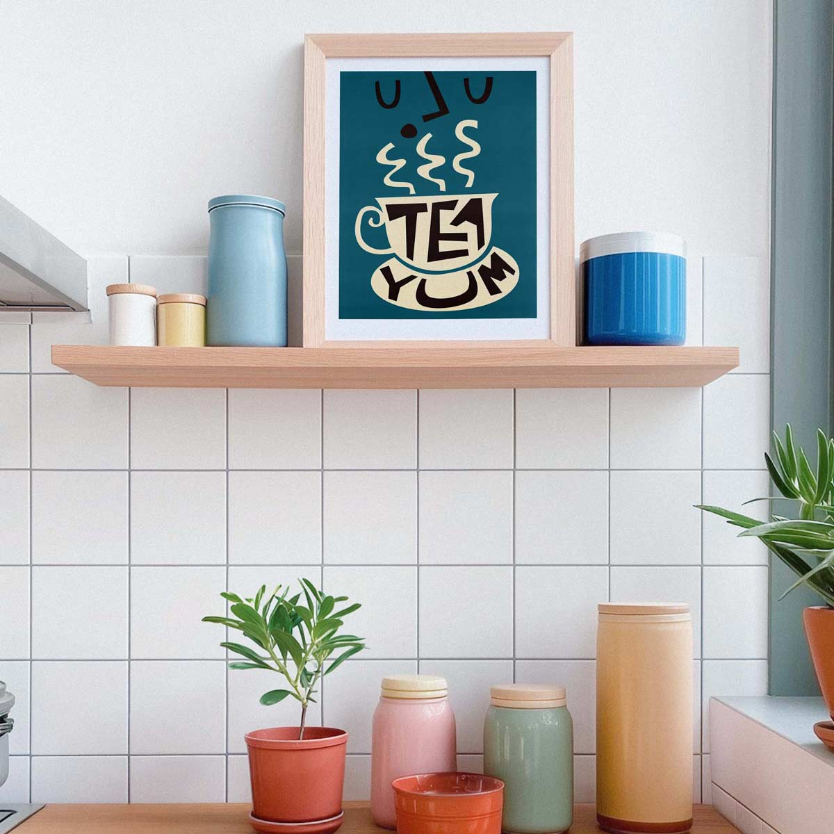 A framed art print with a teal background and a stylized white teacup reading 'TEA YUM' hangs on a light wood shelf in a kitchen setting. The shelf is decorated with blue and cream canisters, and below it, a countertop holds small potted plants and more storage jars against a white tiled wall.