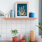 A framed art print with a teal background and a stylized white teacup reading 'TEA YUM' hangs on a light wood shelf in a kitchen setting. The shelf is decorated with blue and cream canisters, and below it, a countertop holds small potted plants and more storage jars against a white tiled wall.
