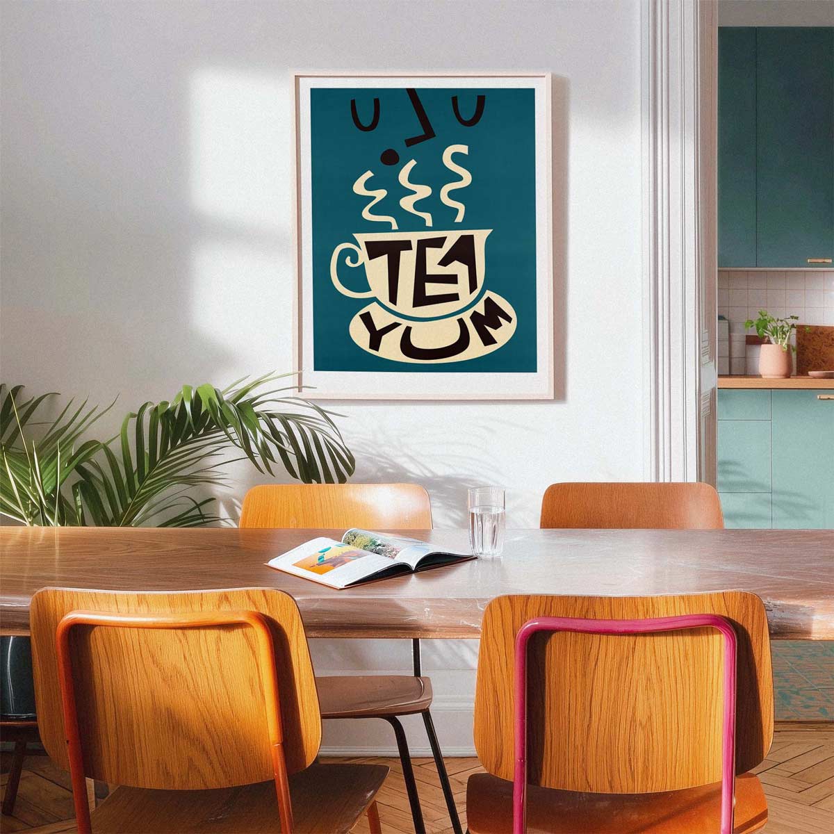 A framed wall art print featuring a stylized teal cup of tea with the words 'TEA YUM' hangs above a wooden dining table with orange chairs in a bright, modern room.